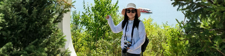 Eine Person posiert mit einer Kamera vor einem malerischen Bergsee, umrahmt von Bäumen und einer modernen Struktur.