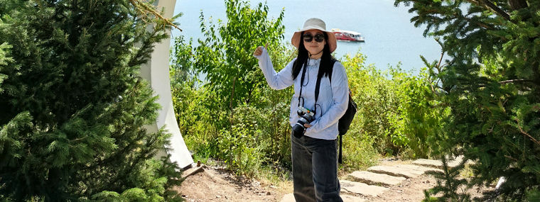 A person stands in a scenic outdoor setting framed by greenery and a modern arch, with mountains and a lake in the background.