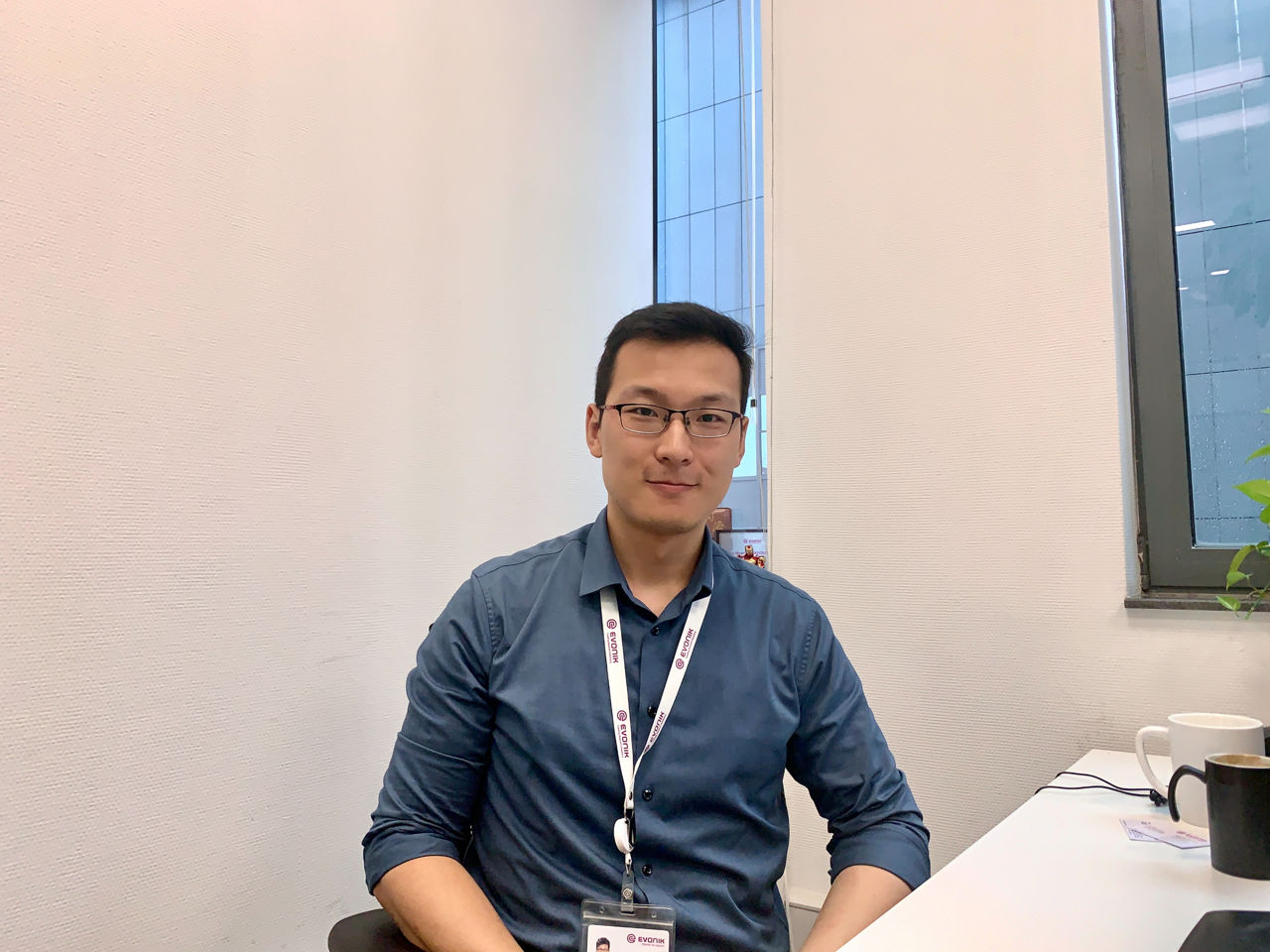 A person sitting at a desk in an office setting, wearing a blue shirt and a lanyard with an ID badge.