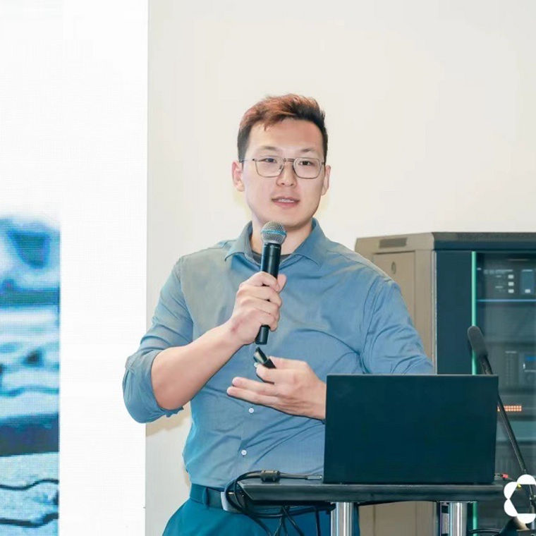 A speaker presents at an event, standing beside a screen displaying abstract visuals.