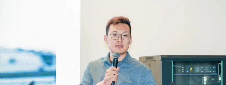 A speaker presents at an event, standing beside a screen displaying abstract visuals.