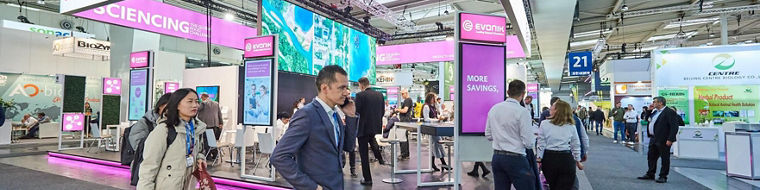Evonik's exhibition booth with bright deep purple lighting and digital screens surrounded by people walking and interacting.
