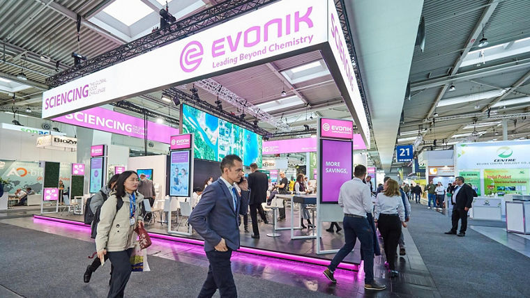 Evonik's exhibition booth with bright deep purple lighting and digital screens surrounded by people walking and interacting.