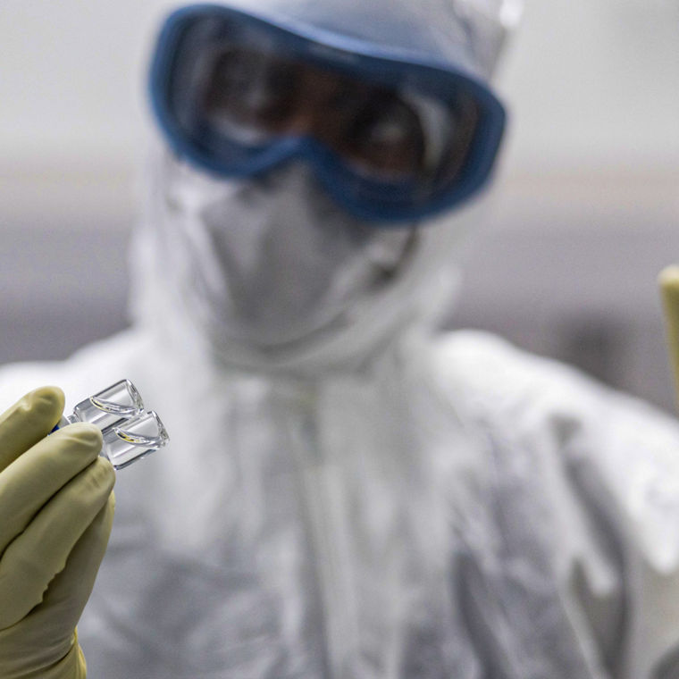 Ein Mann im Vollschutz mit Ganzkörperanzug, Schutzbrille und Gummihandschuhen hält zwei kleine Glasröhrchen.