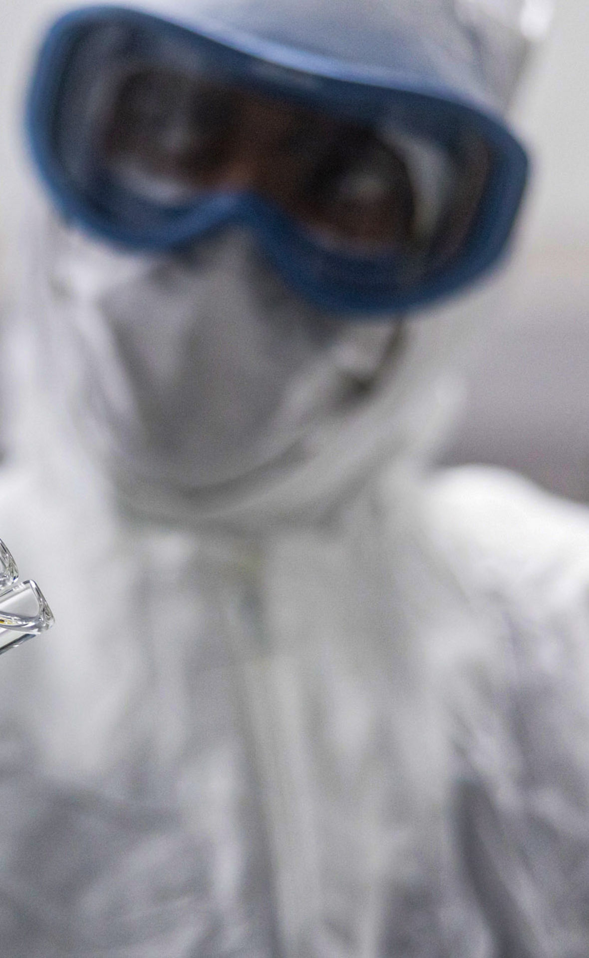 Ein Mann im Vollschutz mit Ganzkörperanzug, Schutzbrille und Gummihandschuhen hält zwei kleine Glasröhrchen.