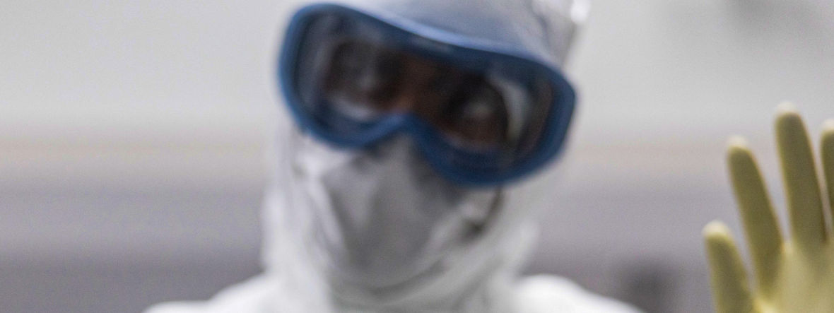 Ein Mann im Vollschutz mit Ganzkörperanzug, Schutzbrille und Gummihandschuhen hält zwei kleine Glasröhrchen.