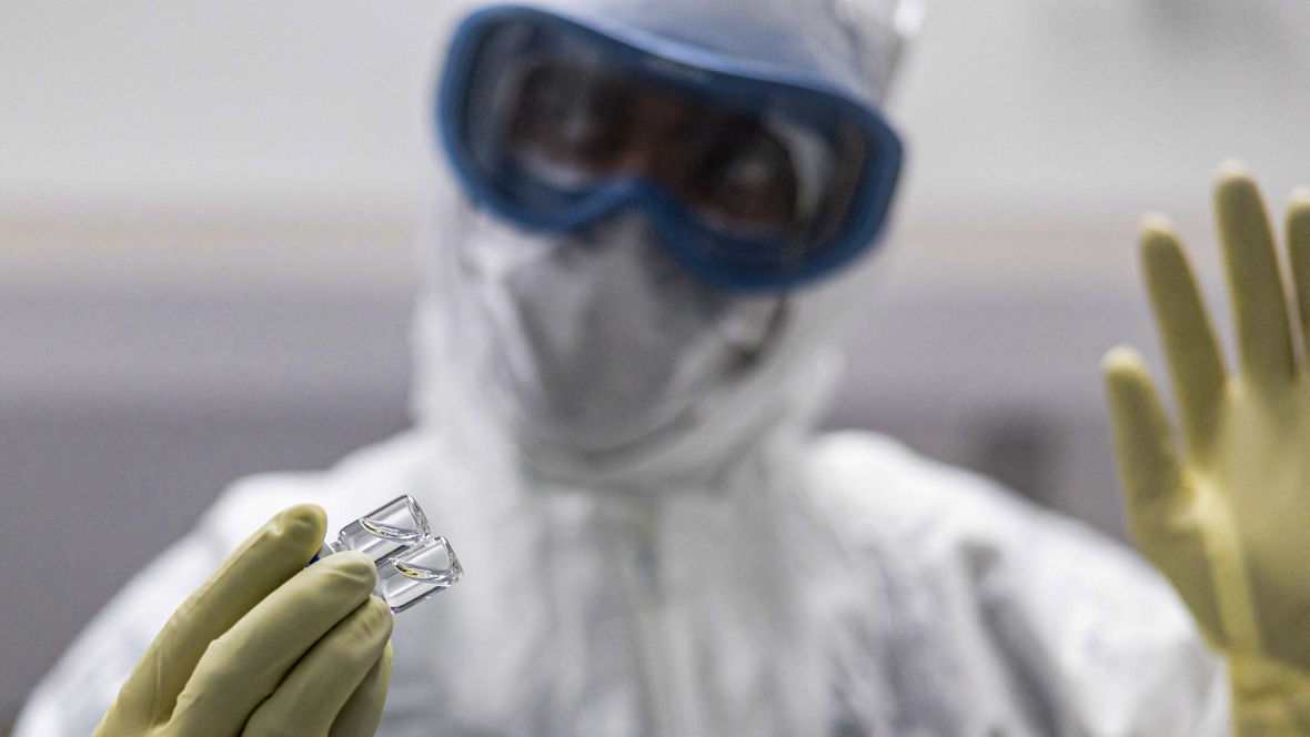 Ein Mann im Vollschutz mit Ganzkörperanzug, Schutzbrille und Gummihandschuhen hält zwei kleine Glasröhrchen.