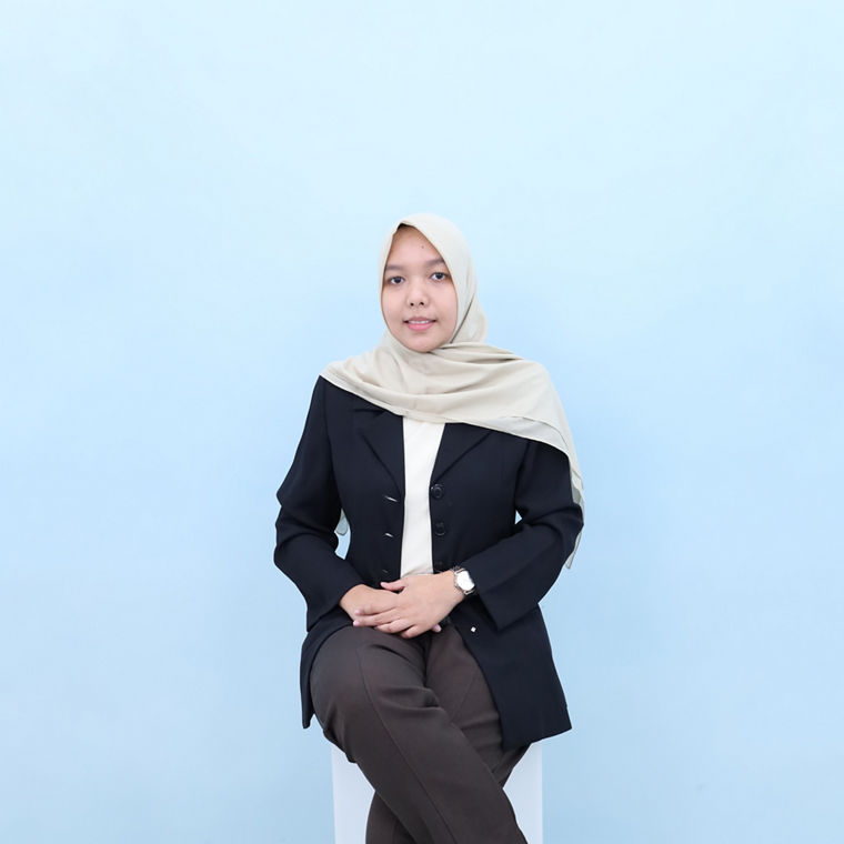 Young woman wearing a beige hijab and black blazer sitting against a light blue background.