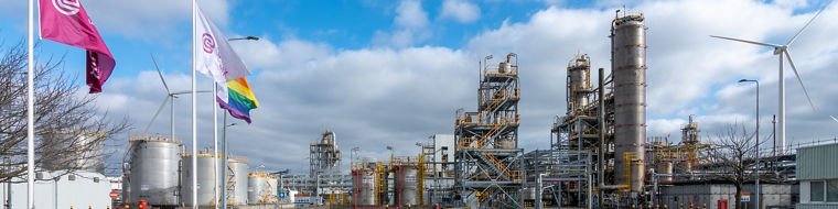 Industrial chemical plant with large storage tanks, metal structures, and wind turbines under a partly cloudy sky.