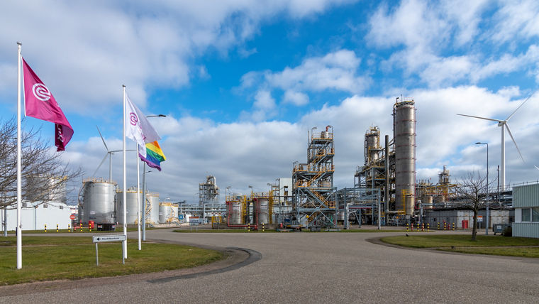 Industrial chemical plant with large storage tanks, metal structures, and wind turbines under a partly cloudy sky.