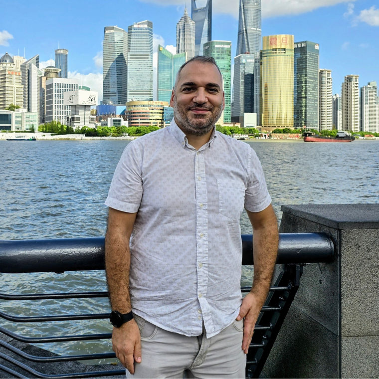 Man standing by a waterfront railing with a city skyline and river in the background under a blue sky.