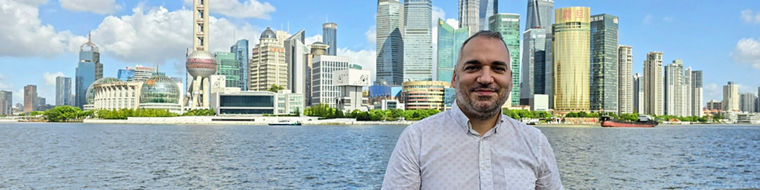 Man standing by a waterfront railing with a city skyline and river in the background under a blue sky.