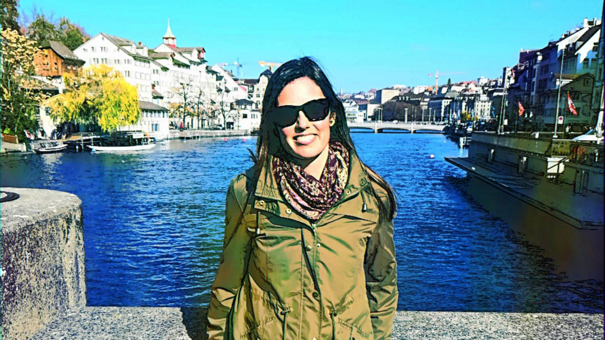 A person stands by the river, wearing a green jacket and jeans, with picturesque buildings and a clear blue sky in the background.