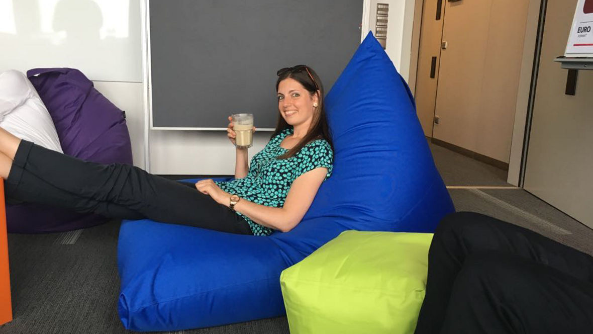 A person relaxes on a blue bean bag while holding a drink, surrounded by colorful bean bags in a casual office setting.