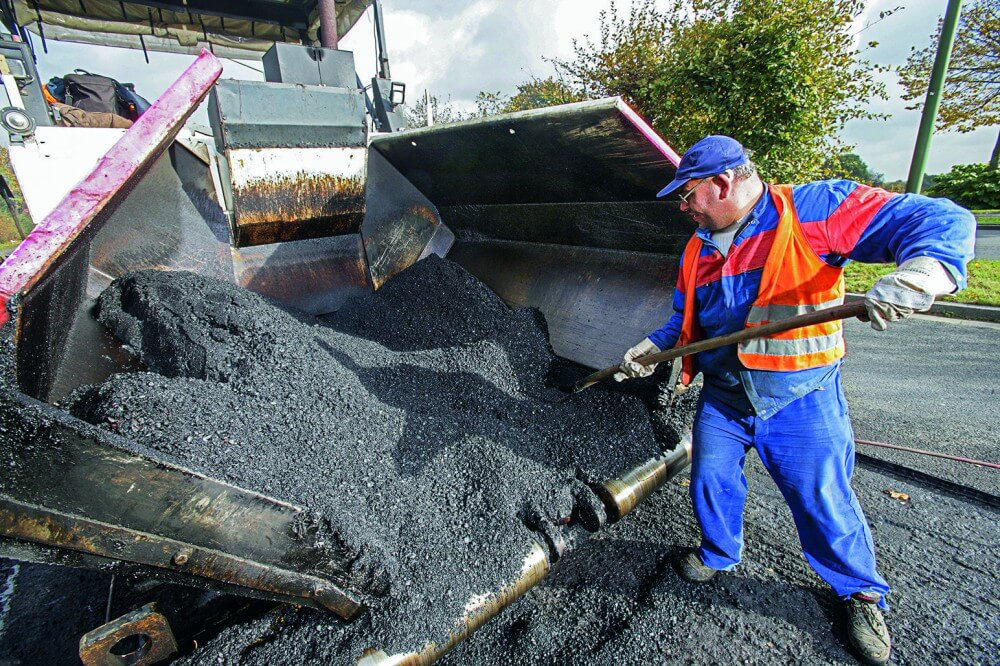 Ein Straßenbauarbeiter verteilt ,it einer Schaufel Roh-Asphalt in einem Straßenbaufahrzeug.