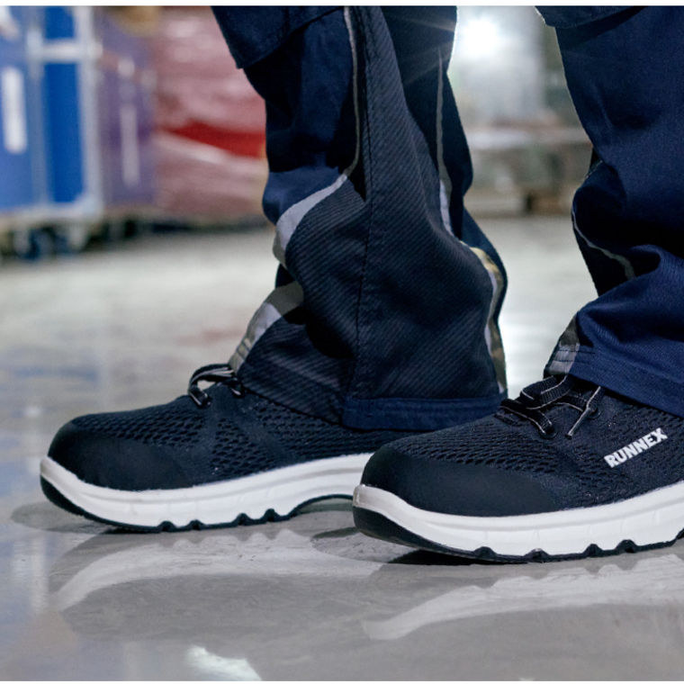 Black and white safety shoes labeled "RUNNEX" worn with dark work pants on a warehouse floor.