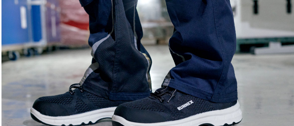 Black and white safety shoes labeled "RUNNEX" worn with dark work pants on a warehouse floor.