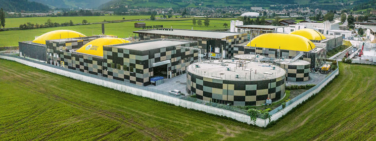 The image shows an aerial view of an industrial facility located in a green, rural area with mountains in the background. The facility includes several large, circular tanks and rectangular buildings. Some of the tanks have yellow domed covers. The buildings and tanks are enclosed by a white fence, and there are grassy fields surrounding the facility. The sky is partly cloudy with sunlight breaking through, illuminating parts of the landscape.