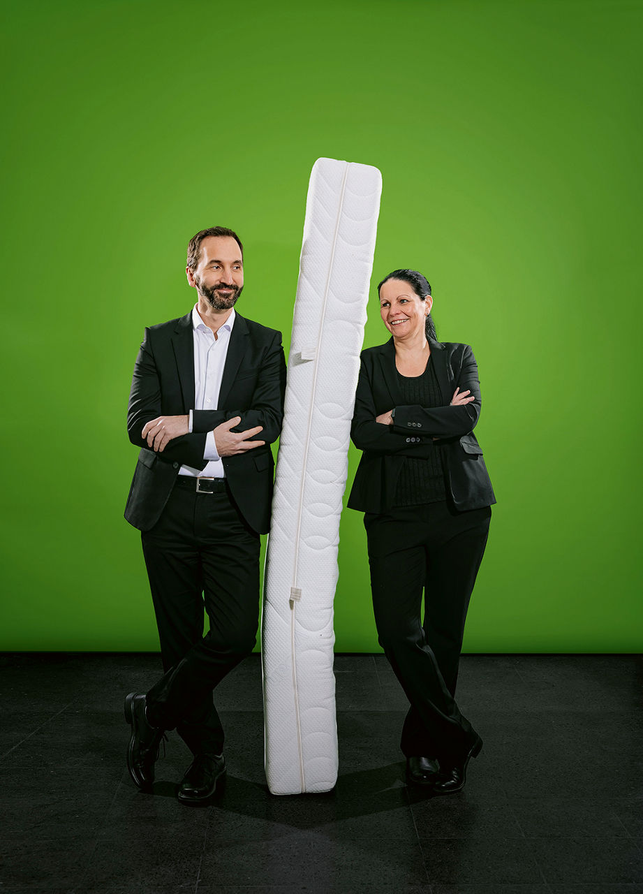 Annegret Terheiden und Michael Ferenz stehen links und rechts neben einer senkrecht aufgestellten Matratze.