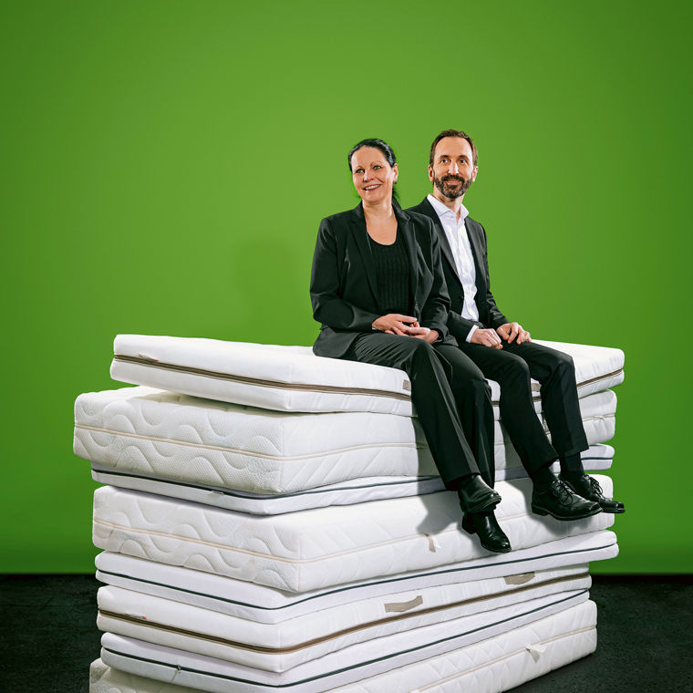 Eine Frau und ein Mann sitzen auf einem Stapel von sechs Matratzen vor einem grünen Hintergrund.