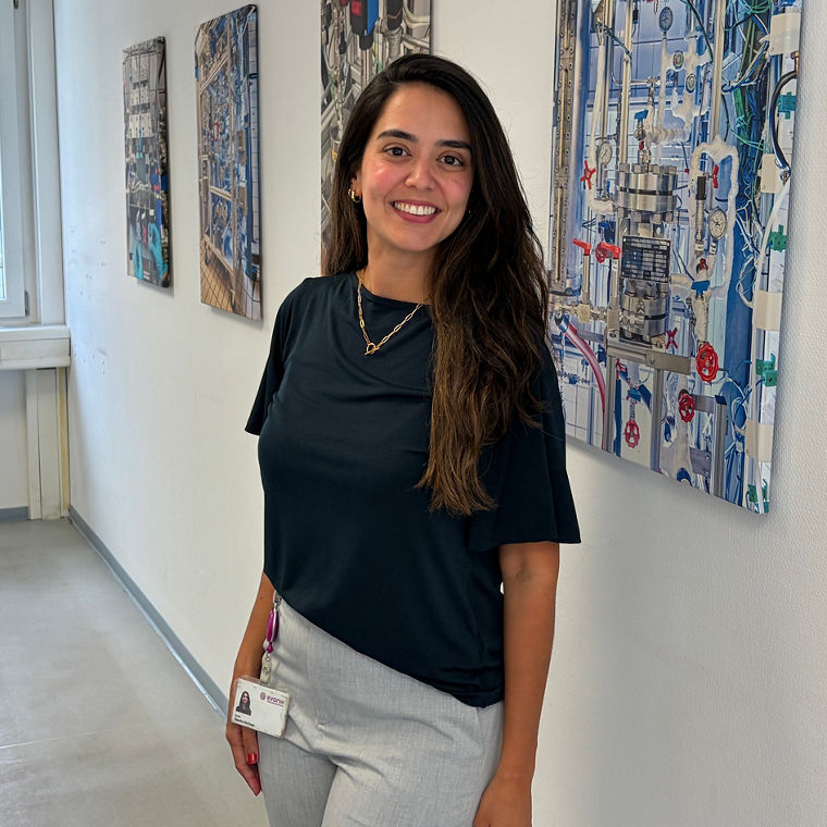 Woman with long dark hair smiling, standing in a hallway with industrial-themed paintings on the wall.