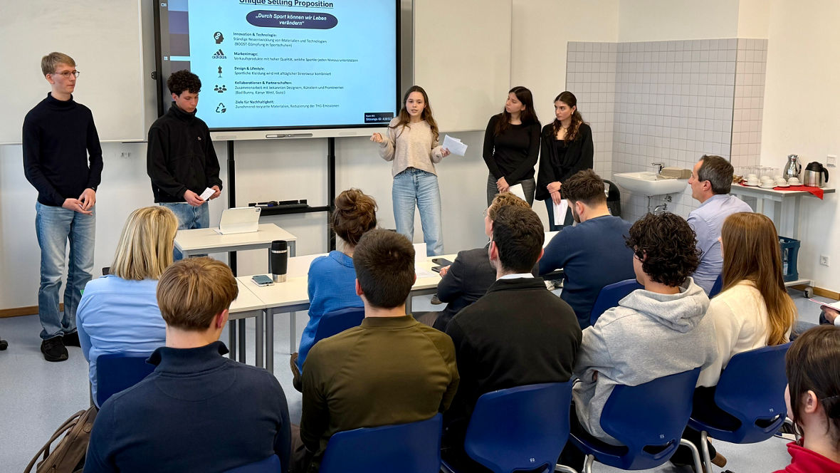 Gruppe von Studierenden präsentiert vor Publikum in einem Klassenraum mit Bildschirm und Flipchart.