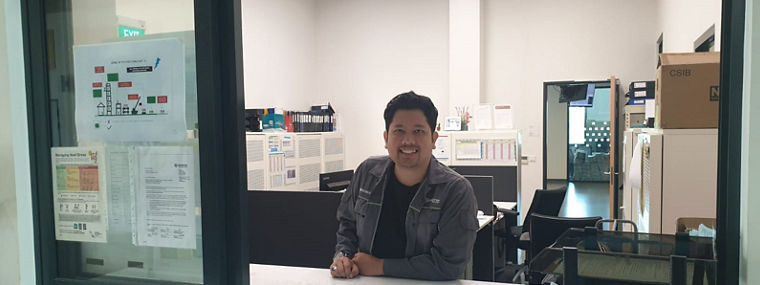 A reception area with a staff member at a service counter and office space in the background.