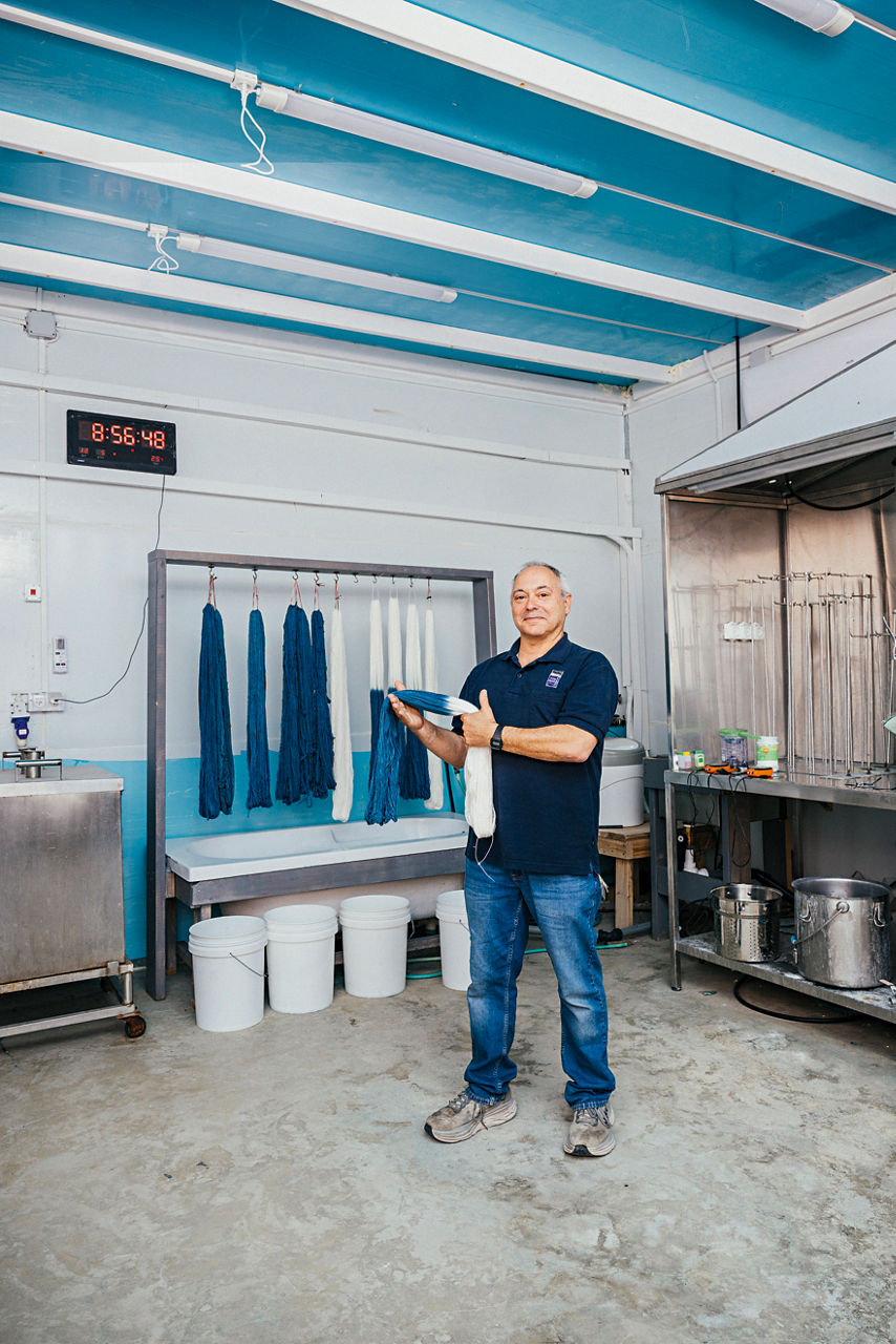 Baruch Sterman steht in der Färberei und hat ein Bündel blau-weißer Gebetsfäden in den Händen. Hinter ihm hängen weitere Bündel.