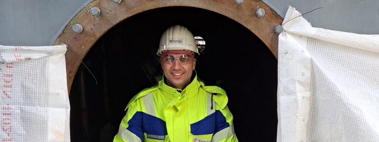 A worker in a high-visibility jacket and gloves sits in an industrial opening, ready for maintenance tasks.