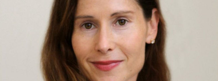 A woman with long hair wearing a white top, smiling and posing against a neutral background.