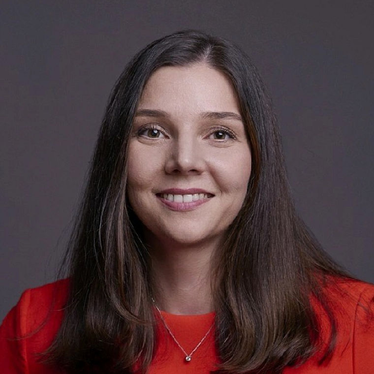 A woman with long hair wearing a red top and a necklace, against a dark background.