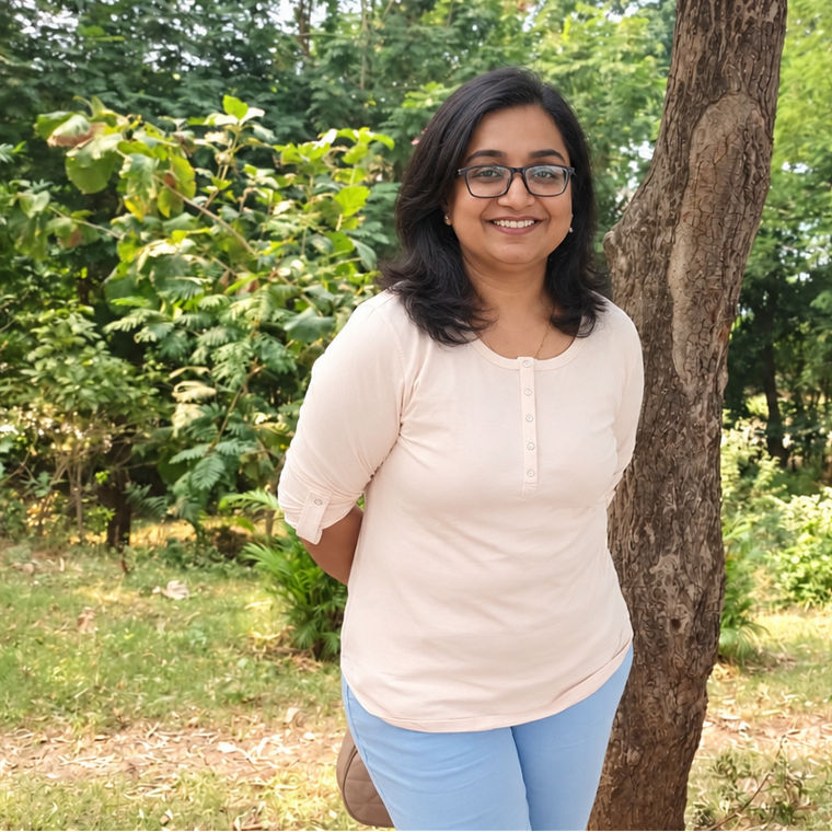 A smiling woman with glasses stands outdoors between two trees in a lush green garden.