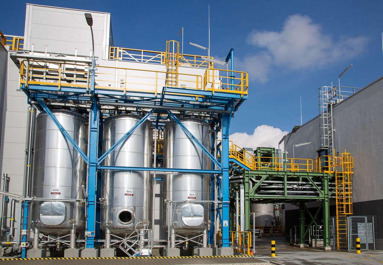 Three steel tanks under a blue sky at Evonik's Slovakian site. A production hall can be seen on the right, the lid of the middle tank is open for inspection.