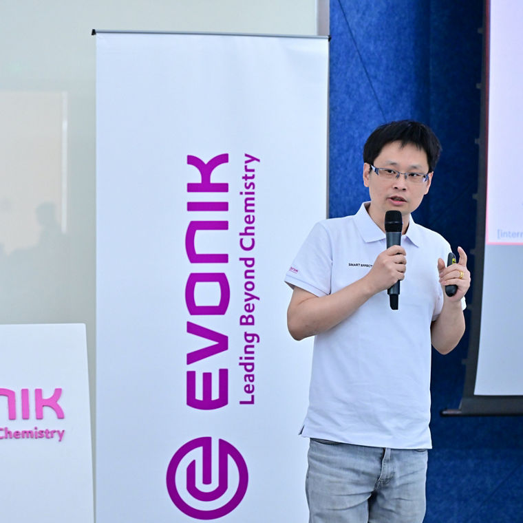A speaker presents at an Evonik event, with the company logo and tagline displayed prominently.