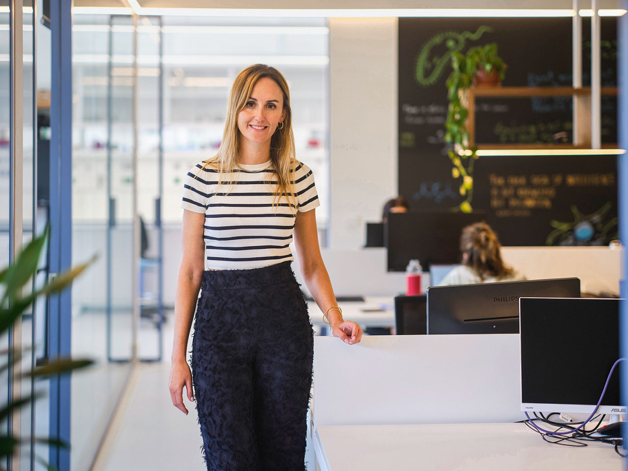 Arta Gil steht neben einem Tisch und schaut in die Kamera. Sie trägt ein gestreiftes Shirt und eine schwarze Hose.