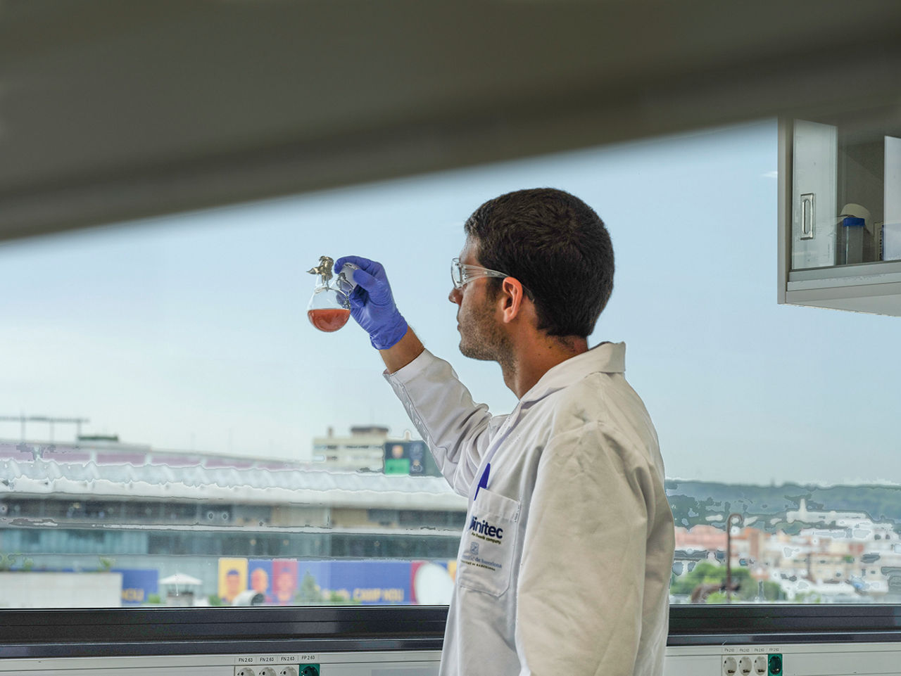 Ein Wissenschaftler steht vor einem Fenster und bewegt ein Kolbenglas mit seiner hand. Draußen ist das Stadiondach von Camp Nou zu erkennen.