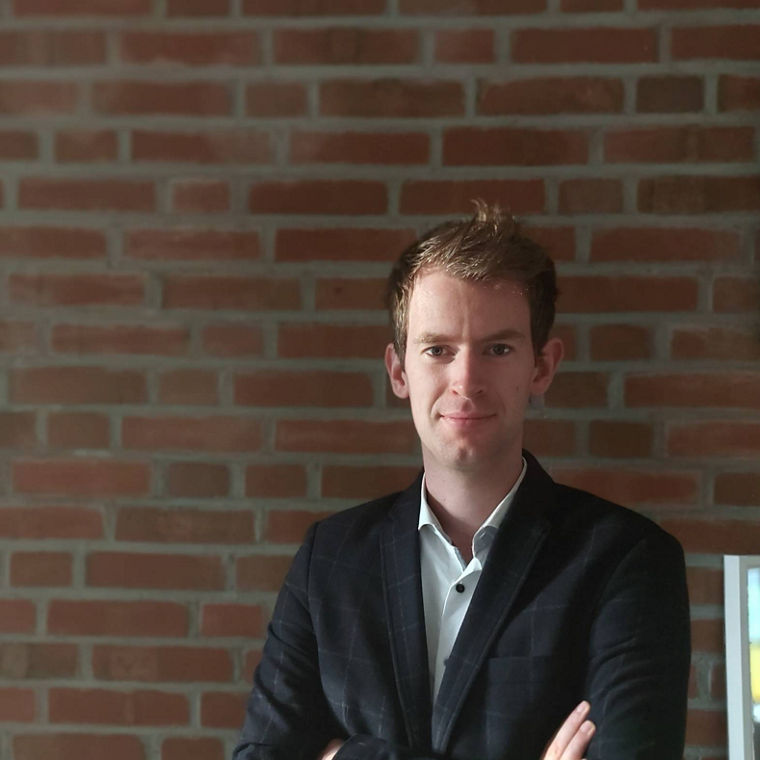 A person in a blazer stands confidently with arms crossed against a brick wall backdrop.