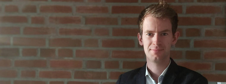 A person in a blazer stands confidently with arms crossed against a brick wall backdrop.