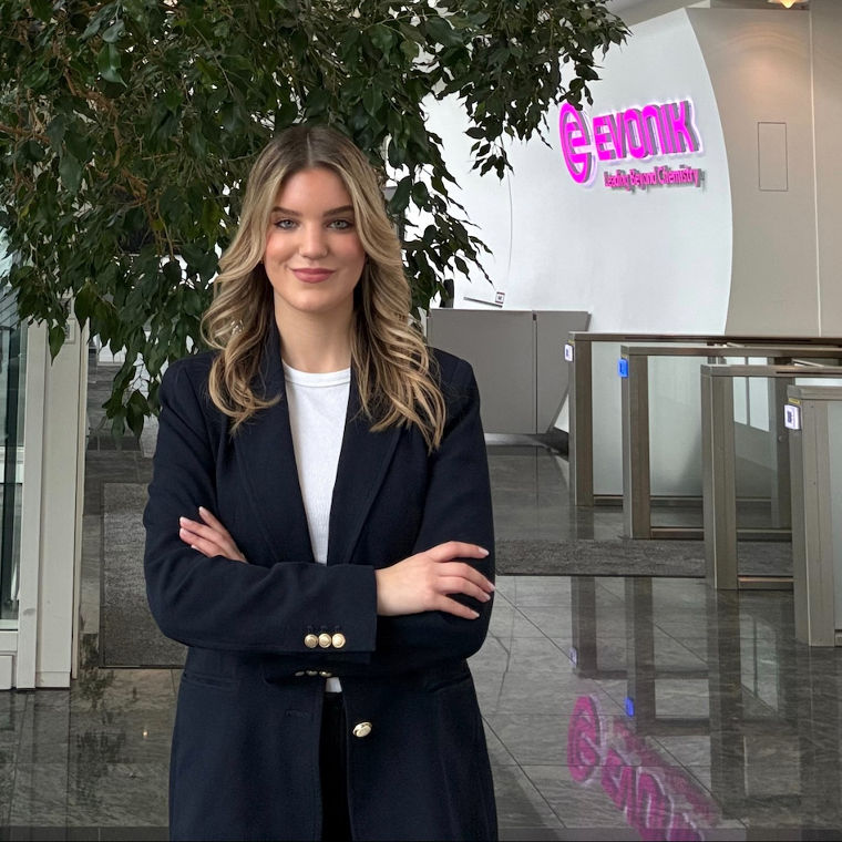 Frau in dunklem Blazer steht vor einem großen Baum im Eingangsbereich mit Evonik-Logo an der Wand.