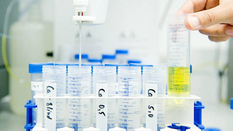 The image depicts a laboratory setting where a person is conducting an experiment involving the measurement of liquid samples. In the foreground, a hand is holding a graduated cylinder filled with a yellow liquid, which is being compared to a row of clear test tubes placed in a rack. Each test tube has a blue lid and is labeled with handwritten identifiers, suggesting they contain different samples or concentrations. A pipette is positioned above one of the test tubes, indicating that a precise amount of liquid is being dispensed. The background features additional laboratory equipment, contributing to the scientific atmosphere of the scene.