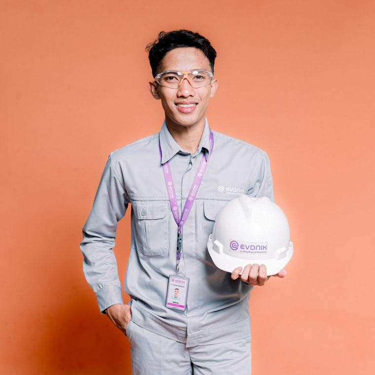 Man in light gray work uniform holding a white safety helmet with a purple logo.