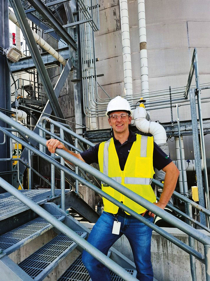 Martin Heining stands on a steel staircase in the facility, wearing a hard hat, safety glasses, and a high-visibility vest, smiling warmly at the camera.