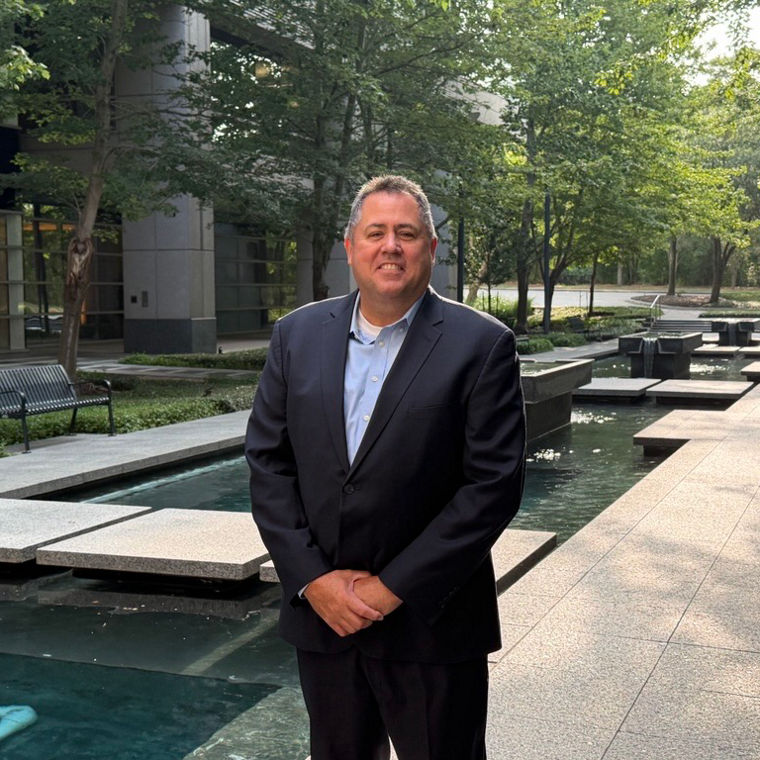Man in a dark suit standing near a modern water feature with trees and benches in the background.