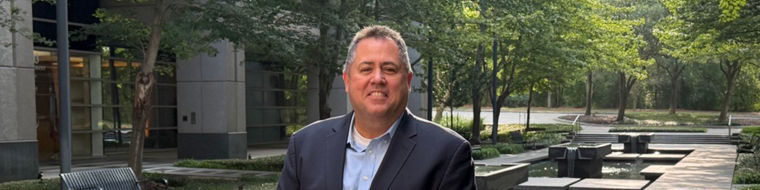 Man in a dark suit standing near a modern water feature with trees and benches in the background.