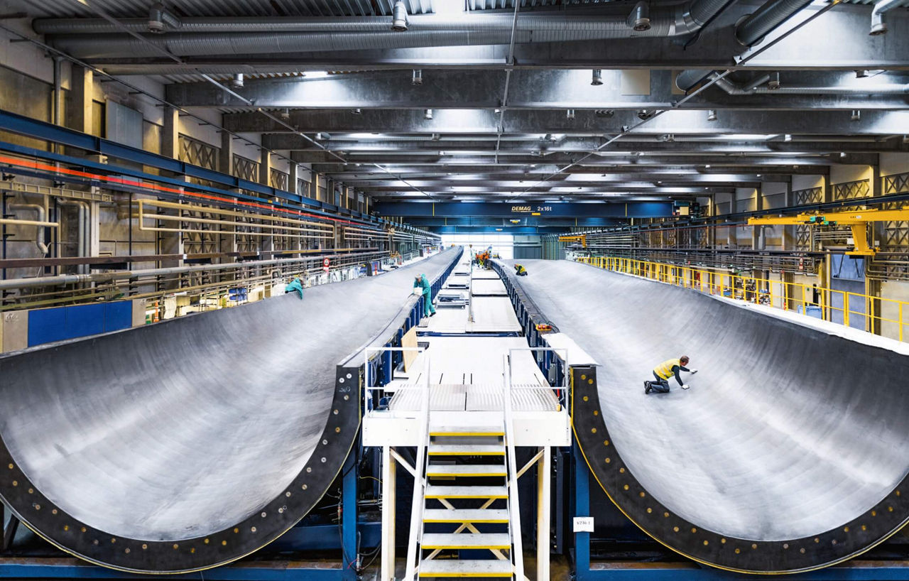 The approximately 100-meter-long molds for rotor blades are stored in a huge hall. People can be seen in the molds preparing them. They look tiny in the surrounding area.