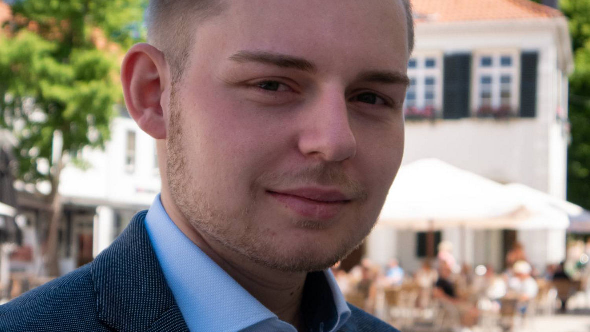 A man in a smart blazer stands outdoors in a lively square with trees and café tables in the background.