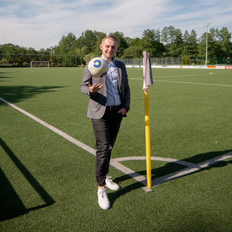 A person in a blazer holds a soccer ball near a corner flag on a sunny sports field.