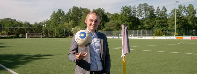 A person in a blazer holds a soccer ball near a corner flag on a sunny sports field.