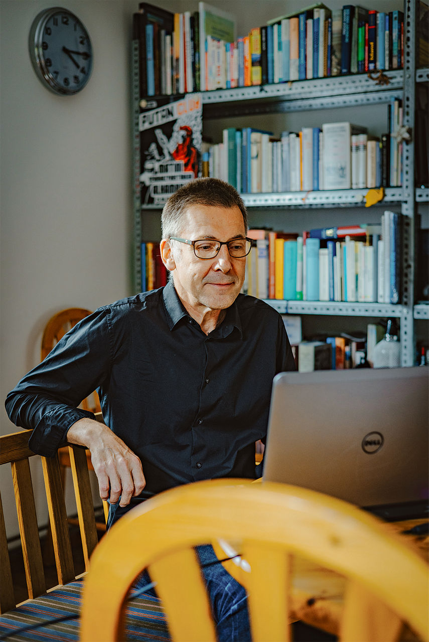 Niko Paech sitzt auf einer Holzbank und nimmt an der Videkonferenz teil. Der Rechner steht auf einem Tisch mit Tischdecke, davor noch ein Stuhl aus Nadelholz.