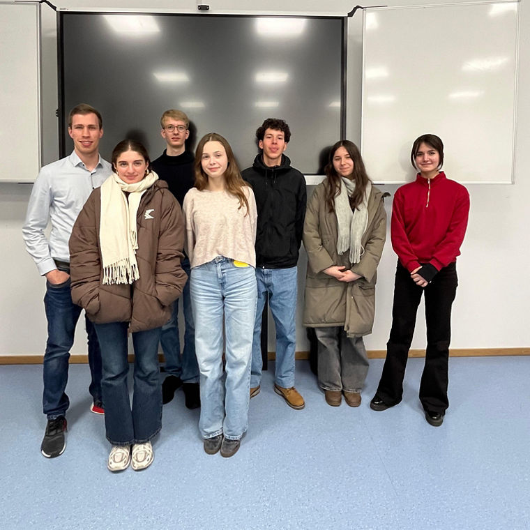 Sieben junge Erwachsene stehen in einem Klassenraum vor einem großen Bildschirm und weißen Tafeln.